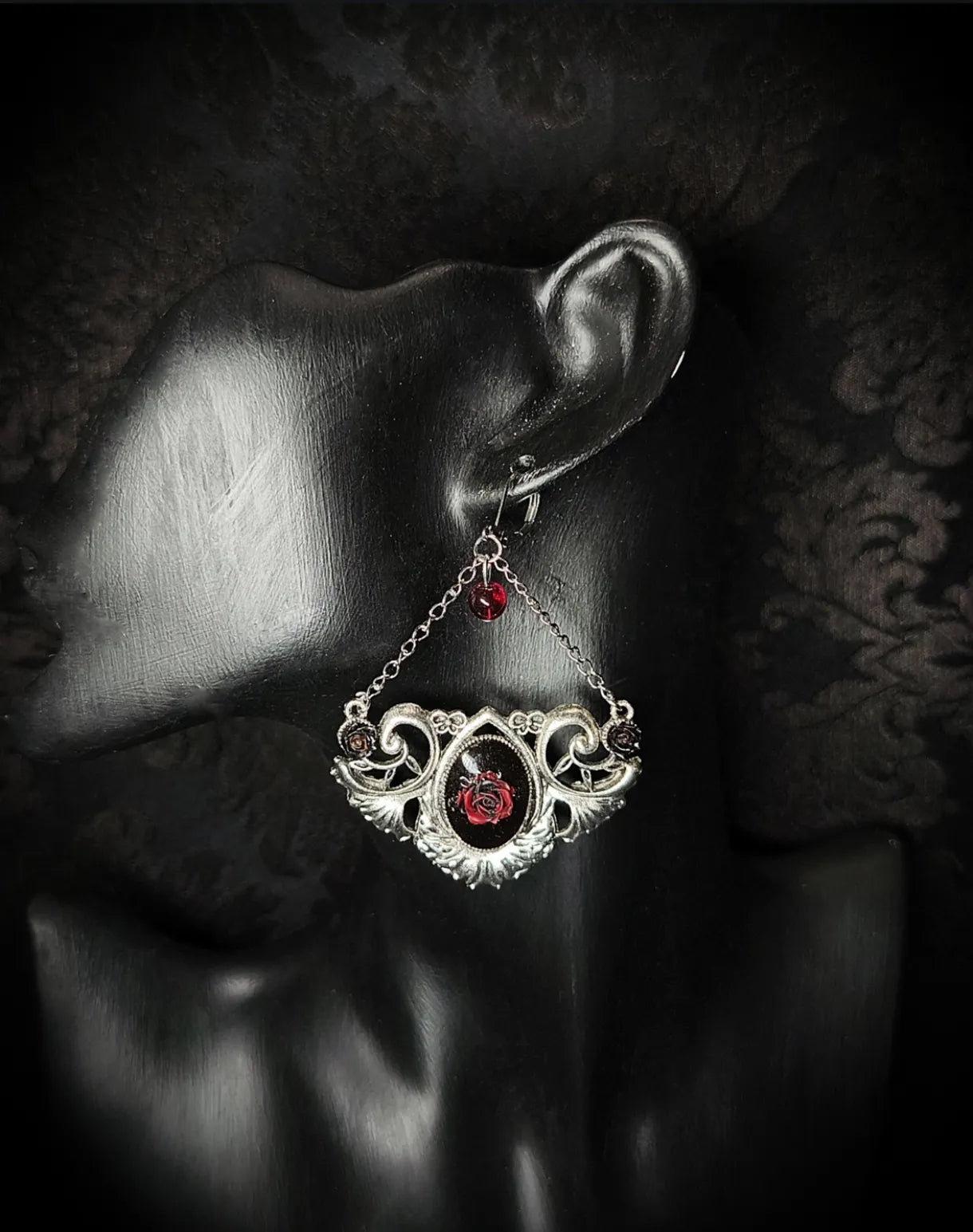 Boucle d’oreille gothique Rosa Nocturna avec rose rouge en résine et ornement argenté portée sur présentoir noir
