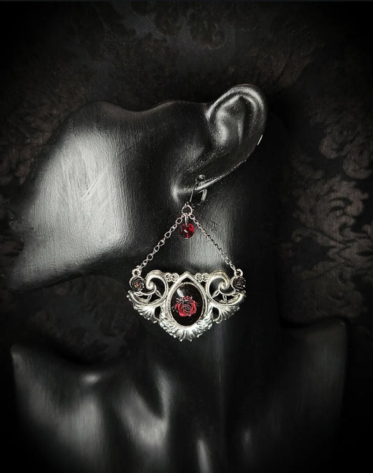 Boucle d’oreille gothique Rosa Nocturna avec rose rouge en résine et ornement argenté portée sur présentoir noir